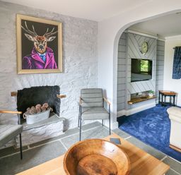 A living room with two chairs near a stone fireplace with pinecones and a deer portrait above and a TV with a clock on the wall at Cwmfadog in Llanafan-fawr near Beulah