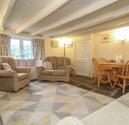 A living room with beige armchairs and sofa a wooden dining table with chairs and a geometric patterned rug at 2 Rose Cottages in Lansallos near Polperro
