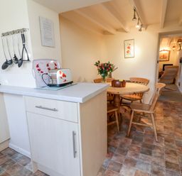 A kitchen and dining area with a round wooden table and chairs and a toaster on the counter at 3 Rose Cottages in Lansallos near Polperro