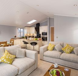 An open living room and kitchen area with beige sofas and yellow cushions at Waternook Lodge South Lakeland Leisure Village