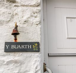 A white textured wall with a rusted metal bell and a sign reading Y Buarth next to a white paneled door at Y Buarth in Waunfawr