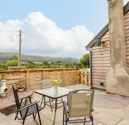 An outdoor patio with a glass table and four chairs next to a wooden fence and a building wall with a chimney at Bryn Hyfryd in Harlech