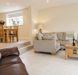 A living room with a beige sectional sofa a wooden coffee table with wine bottles glasses and a bowl a wooden dining table with chairs and flowers on it at Hen Dy Bryn Mawndy Melin y Wig near Corwen