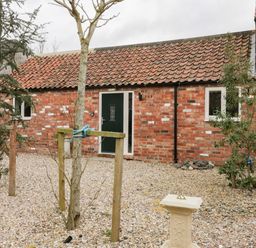 A gravel courtyard with small trees and a sundial in front of a brick building with a tiled roof and green door at Robins Nest Cottage in Brandesburton