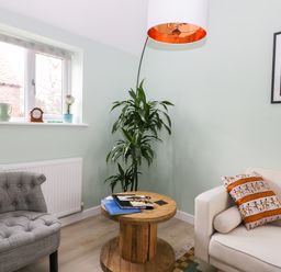 A small living room with a gray armchair a white sofa a wooden round coffee table with books a tall plant a window with decorations and a hanging lamp at Robins Nest Cottage in Brandesburton