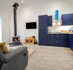 A living area with a dark blue kitchen, wood-burning stove, TV on the wall, and gray armchair at 3 Mountain View in Talwrn near Llangefni