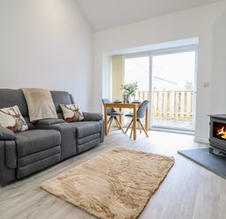 A living room with a grey sofa, a small wooden dining table with two chairs, a beige rug, and a wood burning stove at 3 Mountain View in Talwrn near Llangefni