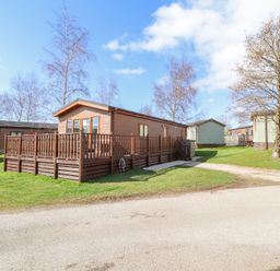 An exterior view of a wooden lodge with a deck surrounded by grass and trees at South Lakeland Leisure Village