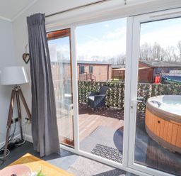 A room with a floor lamp and a wooden table next to glass sliding doors leading to a deck with a chair and a hot tub at Duckling Lodge in South Lakeland Leisure Village