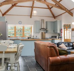 An open plan kitchen and living area with wooden beams dining table and chairs a brown leather sofa a black sofa and kitchen cabinets at The Old Haybarn in Ipplepen