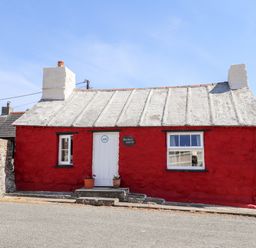 A small red stone cottage with a white door and two windows on a street at Bwthyn Alarch in Trefin St Davids Pembrokeshire