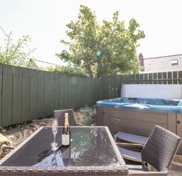 An outdoor patio with a glass table and wicker chairs a bottle of wine two glasses a green wooden fence and a hot tub at Bwthyn Alarch in Trefin St Davids Pembrokeshire