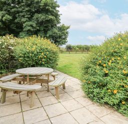 An outdoor patio area with a round wooden picnic table and benches surrounded by flowering shrubs and a grassy field at The Granary in Newport Pembrokeshire