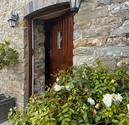 An open wooden door in a stone wall with plants and white flowers outside at The Granary in Newport Pembrokeshire