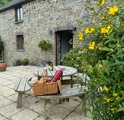 An outdoor stone patio with a round wooden picnic table and benches with a basket a bottle and glasses near a stone building with potted plants and yellow flowers at The Granary in Newport Pembrokeshire