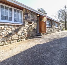 Stone cottage exterior with three white framed windows and an open wooden door on a paved path at Otter Cottage in Dinas Cross