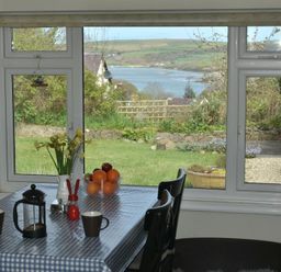 A dining area with a table covered by a checkered cloth and chairs by a large window with a view of a garden and water at Gellifor in St Dogmaels Pembrokeshire