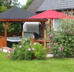 A garden with pink flowers a wooden gazebo with a hot tub a red umbrella and a stone house at Pond Cottage in New Quay Ceredigion