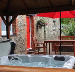 An outdoor patio area with a hot tub a wooden table and chairs with a red umbrella attached to the table next to a stone wall with a red door and window at Pond Cottage in New Quay Ceredigion