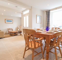 A dining area with a wooden table set with white dishes and glasses next to a window with curtains and an adjacent living room with sofas and wicker furniture at Neverndale in Newport Pembrokeshire