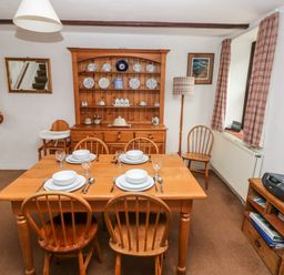 A dining room with a wooden table set for four people wooden chairs a wooden cabinet with plates and jars a floor lamp and a small shelf with a radio at Ty Canol in Mathry Pembrokeshire