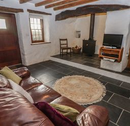 A living room with a leather sofa and cushions a wood burning stove a small round rug a wooden chair and a television on a wall shelf at Panteurig Cottage Strumble Head Goodwick Pembrokeshire