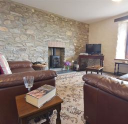 A living room with brown leather sofas a stone wall with a fireplace a wooden TV stand with a television and a window at Panteurig Farmhouse in Strumble Head Goodwick Pembrokeshire