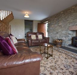 A living room with brown leather sofas a small wooden table a stone wall fireplace a basket of firewood and a floral carpet at Panteurig Farmhouse in Strumble Head Goodwick Pembrokeshire