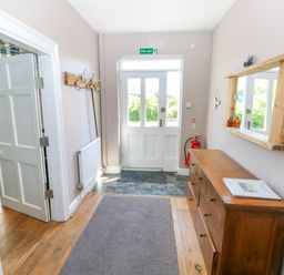 An entrance hallway with a wooden dresser and mirror on the right wall a coat rack on the left wall and a white door with a fire exit sign above at Newton West Farm Newton near Newgale