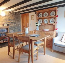 A dining area with a wooden table and chairs a wooden cabinet with decorative plates a wooden bookshelf with books a gray armchair with a cushion and a stone wall at Gwndwn Gwyn in Trefasser Near Strumble Head