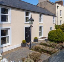 Front exterior of a beige house with six white-framed windows a black door a black lamp post two potted plants and garden shrubs at The Rock House in Fishguard Pembrokeshire