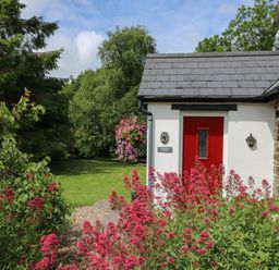 A cottage entrance with a red door surrounded by pink flowers and green trees at Trawsnant Cottage in New Quay Ceredigion