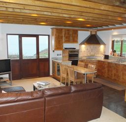 A kitchen and living area with a brown leather sofa wooden dining chairs a television and wooden cabinets at Cable Cottage in Fishguard Pembrokeshire