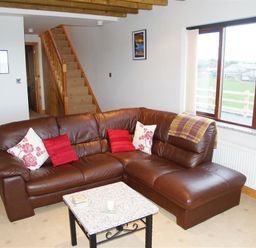A living room with a brown leather sectional sofa with cushions a small table stairs and window with a view at Cable Cottage in Fishguard Pembrokeshire