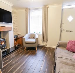 A living room with a wood stove fireplace armchairs a sofa with cushions a wall-mounted television and a front door at Scarborough Cottage in Driffield
