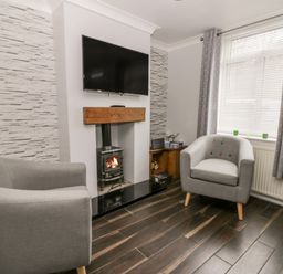 A living room with two grey armchairs a wood burning stove under a wall mounted tv and a window with blinds and curtains at Scarborough Cottage in Driffield