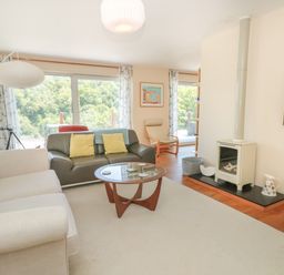 A living room with sofas a glass coffee table a wood stove and large windows showing trees outside at High Banks in Dartmouth