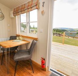A small dining area with a wooden table and two chairs by a window with a striped blind and a door opening to a deck and grassy outdoor area at Marches Way in Rhayader