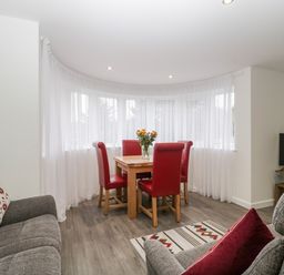 A dining area with a wooden table and red chairs next to curved windows with white curtains and a living space with grey sofas and a television at Flat 2 38 Preston Road in Weymouth