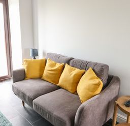 A gray sofa with four yellow cushions next to a small wooden table with a blue lamp at Tyddyn Haidd in Amlwch