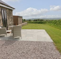 A stone building with a patio table and two chairs on a stone slab area next to a gravel path and lawn at Tyddyn Haidd in Amlwch