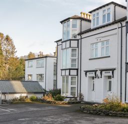 The exterior of a white multi-story building with large windows a small garage and a parking area surrounded by trees at Tethera in Bowness-On-Windermere
