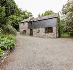 A gravel driveway leading to a stone house with a slate roof surrounded by trees and plants at Ribby Barn in Lerryn