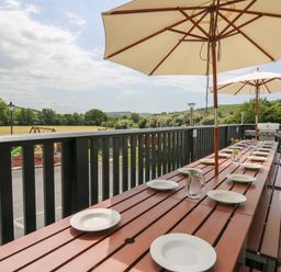 An outdoor dining table with plates glasses jug and umbrellas on a wooden deck overlooking a field with trees at Llety Ceiro in Bow Street Wales