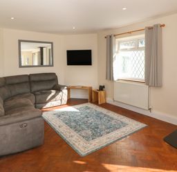 A living room with a gray sectional sofa a blue and white rug a wall-mounted TV a window with gray curtains and a wood-burning stove at Ty Garth in Benllech