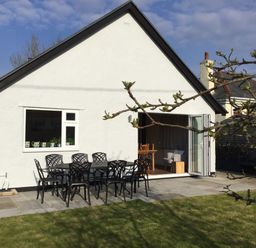 A white house with a patio table and chairs on a stone terrace and a grassy yard at Ty Garth in Benllech