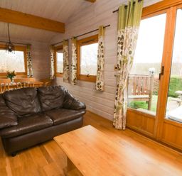 A room with a brown leather sofa a wooden coffee table and a dining table with chairs near windows at Barn Owl Lodge Rhayader