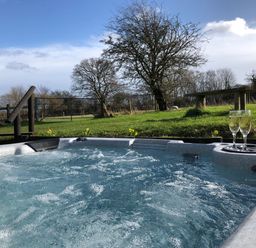 An outdoor hot tub with two glasses of champagne on a tray overlooking a grassy area with trees and a wooden bench at Deer Lodge in Wimborne Minster