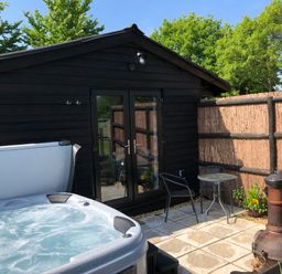 A patio area with a hot tub table with two chairs a chiminea a black wooden building and a fence at Deer Lodge in Wimborne Minster
