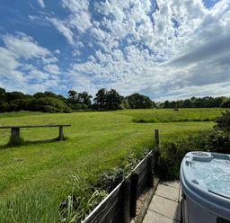 An outdoor area with a hot tub patio bench and a large grassy field with trees under a blue sky with clouds at Deer Lodge in Wimborne Minster